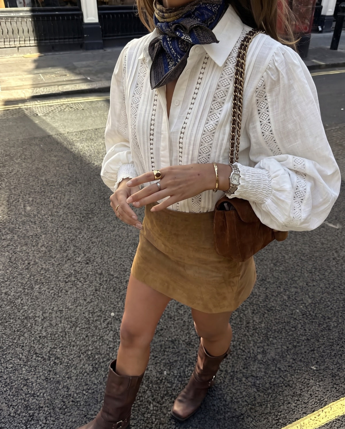 Blusa Cefalù branca em 100% algodão, com aplicações de renda e pregas frontais, combinada com minissaia castanha, botas de cano alto e lenço estampado.