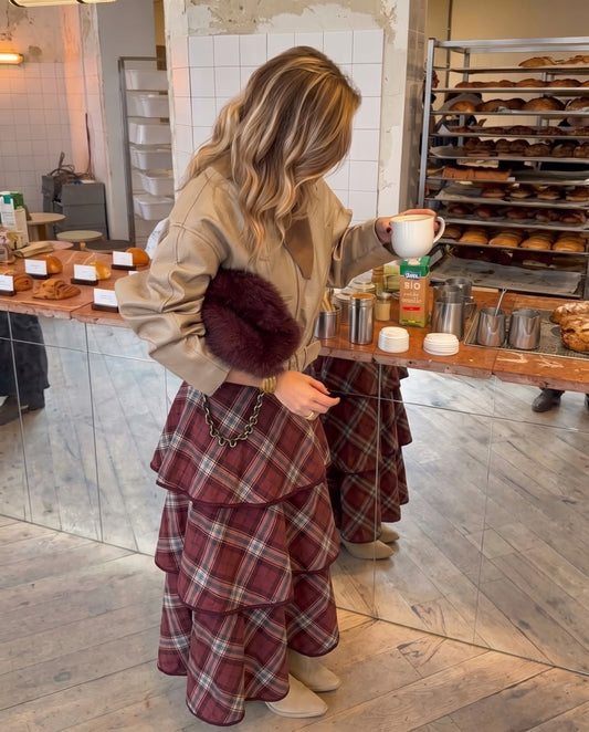 Saia San Gimignano bordô com padrão xadrez e quatro camadas de folhos, combinada com casaco bege e botas, num look feminino de outono.
