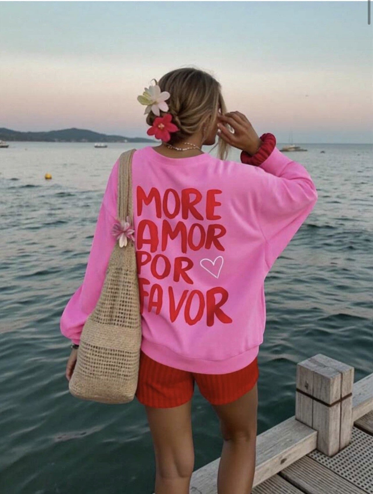 Shevibes sweatshirt rosa choque com "MORE AMOR POR FAVOR" em letras vermelhas e um pequeno coração branco. Modelo com flores no cabelo.